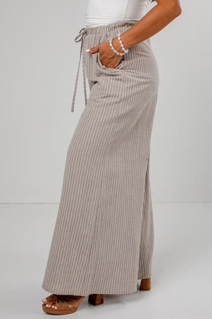 A person wearing a high-waisted, wide-leg maxi skirt with vertical stripes in beige and brown, paired with a white top and brown platform sandals, shown from the waist down against a light gray studio background.