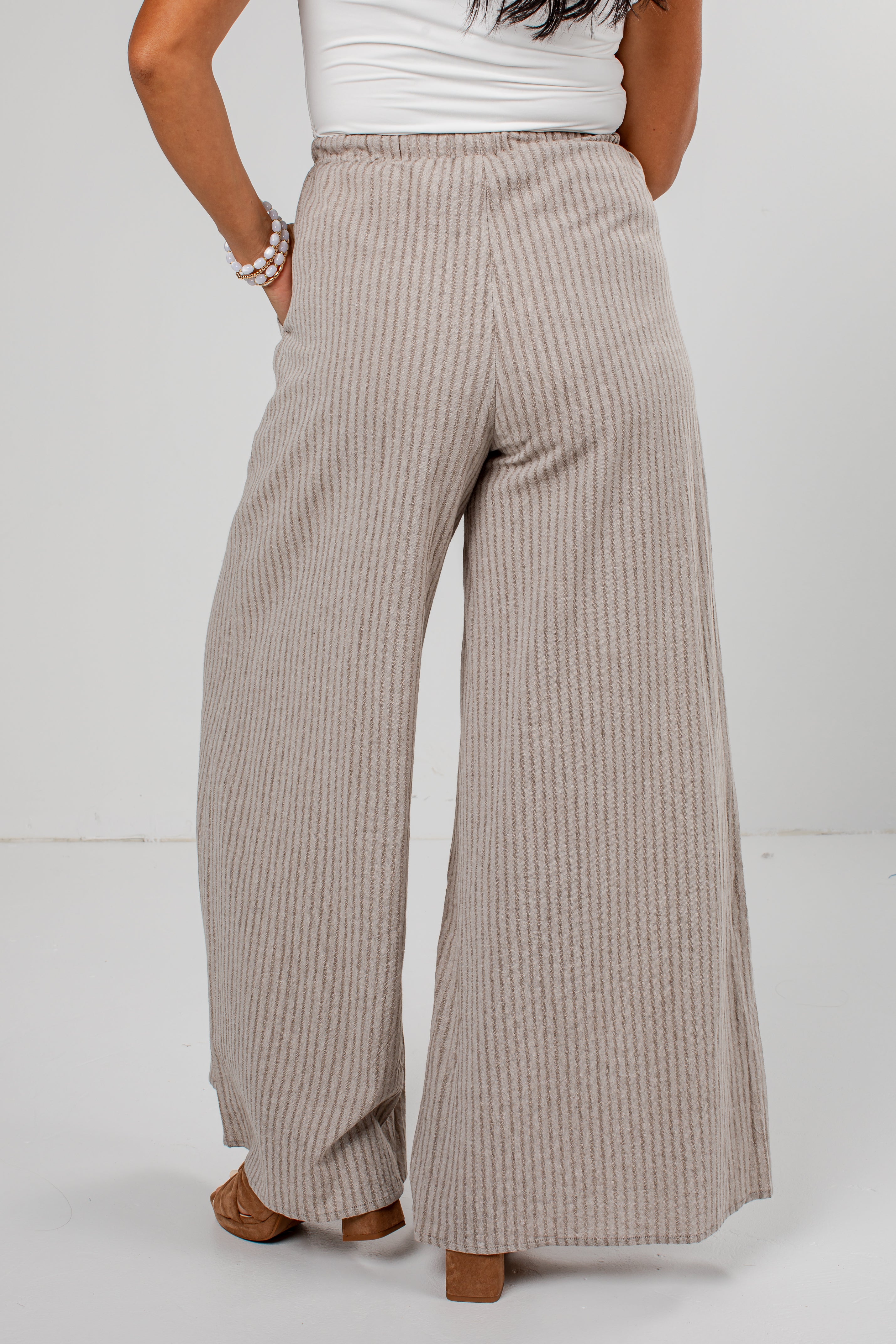 A person shown from the back wearing high-waisted, wide-leg pleated pants in a light beige and white stripe pattern, paired with a white top and brown block-heel sandals.
