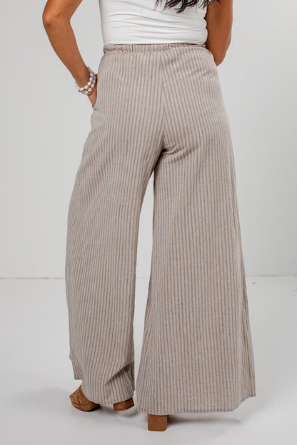 A person shown from the back wearing high-waisted, wide-leg pleated pants in a light beige and white stripe pattern, paired with a white top and brown block-heel sandals.