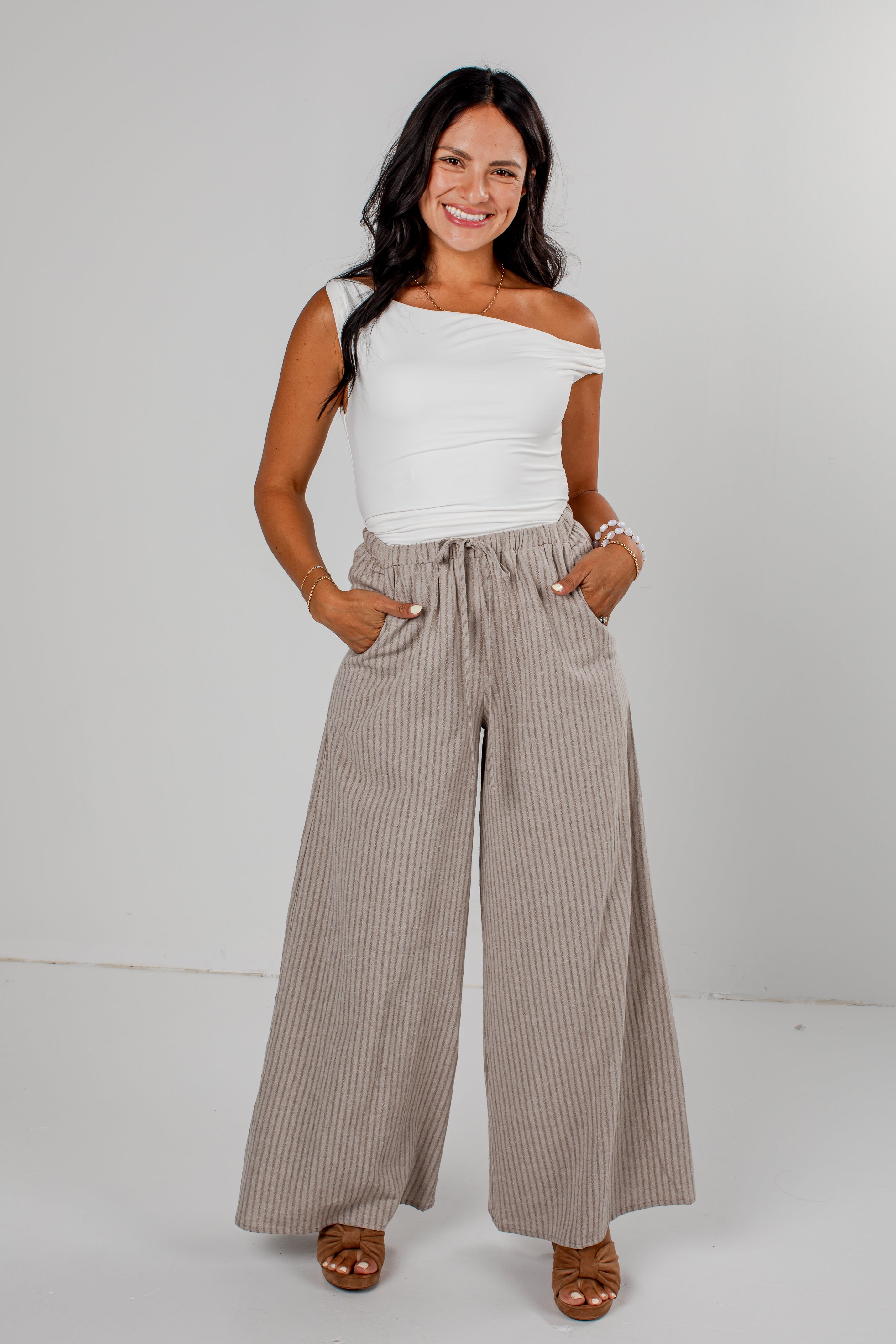 A woman modeling a white off-the-shoulder top and high-waisted, wide-leg taupe pinstripe pants against a light gray studio background.