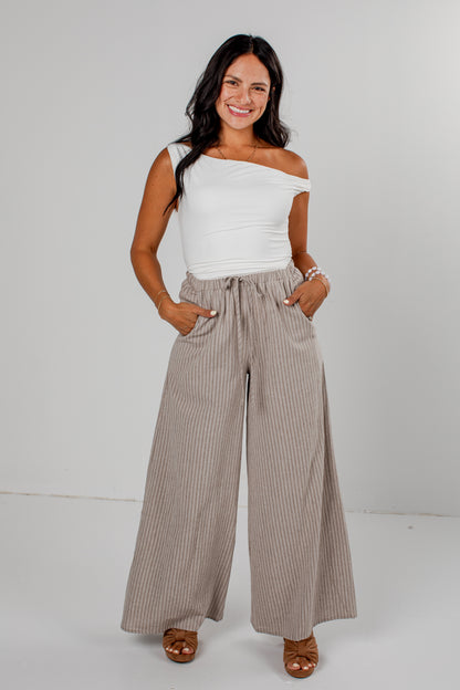 A woman modeling a white off-the-shoulder top and high-waisted, wide-leg taupe pinstripe pants against a light gray studio background.