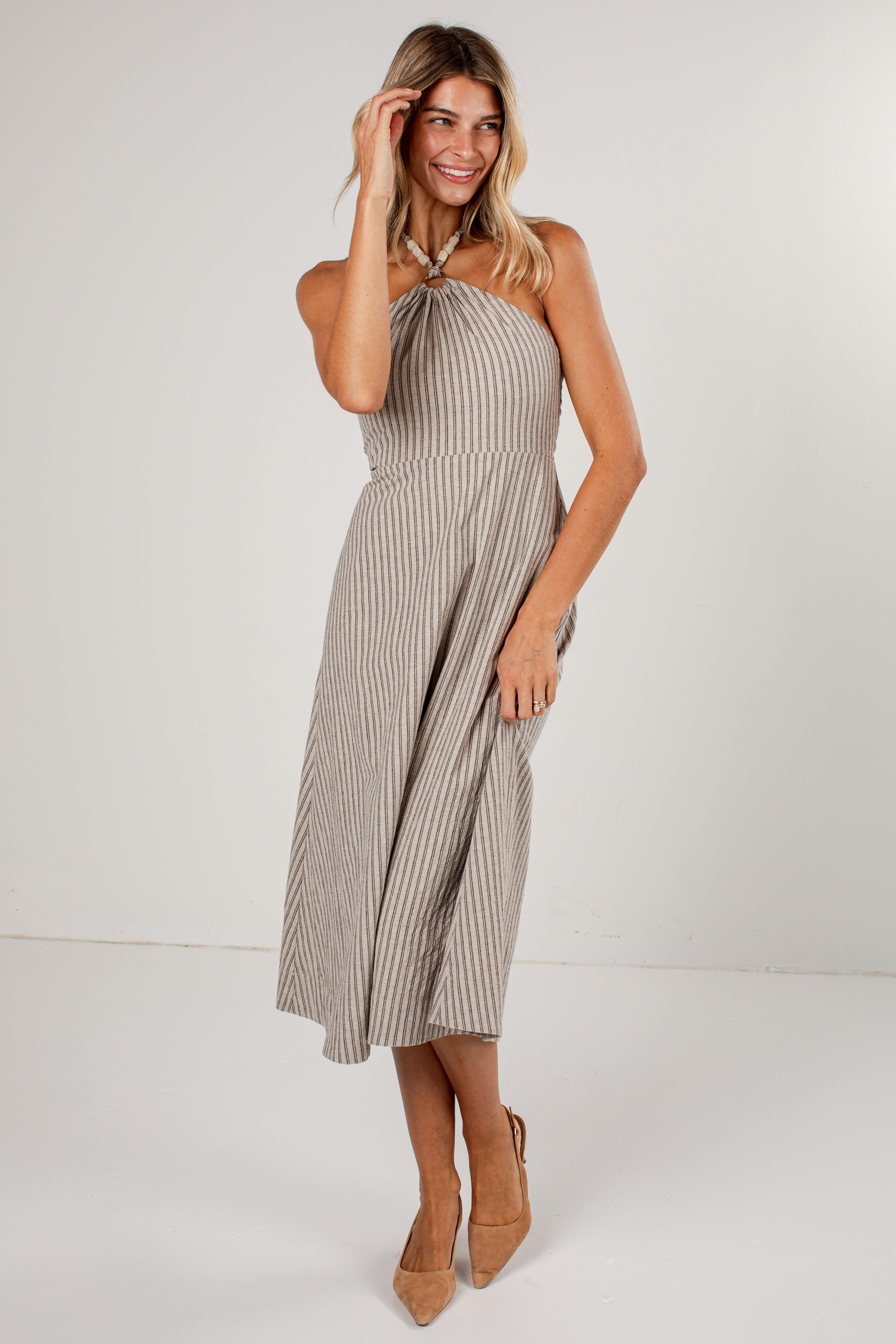A woman modeling a sleeveless, halter-style midi dress with vertical pinstripes in beige and white, paired with tan high heels, against a plain light gray studio background.