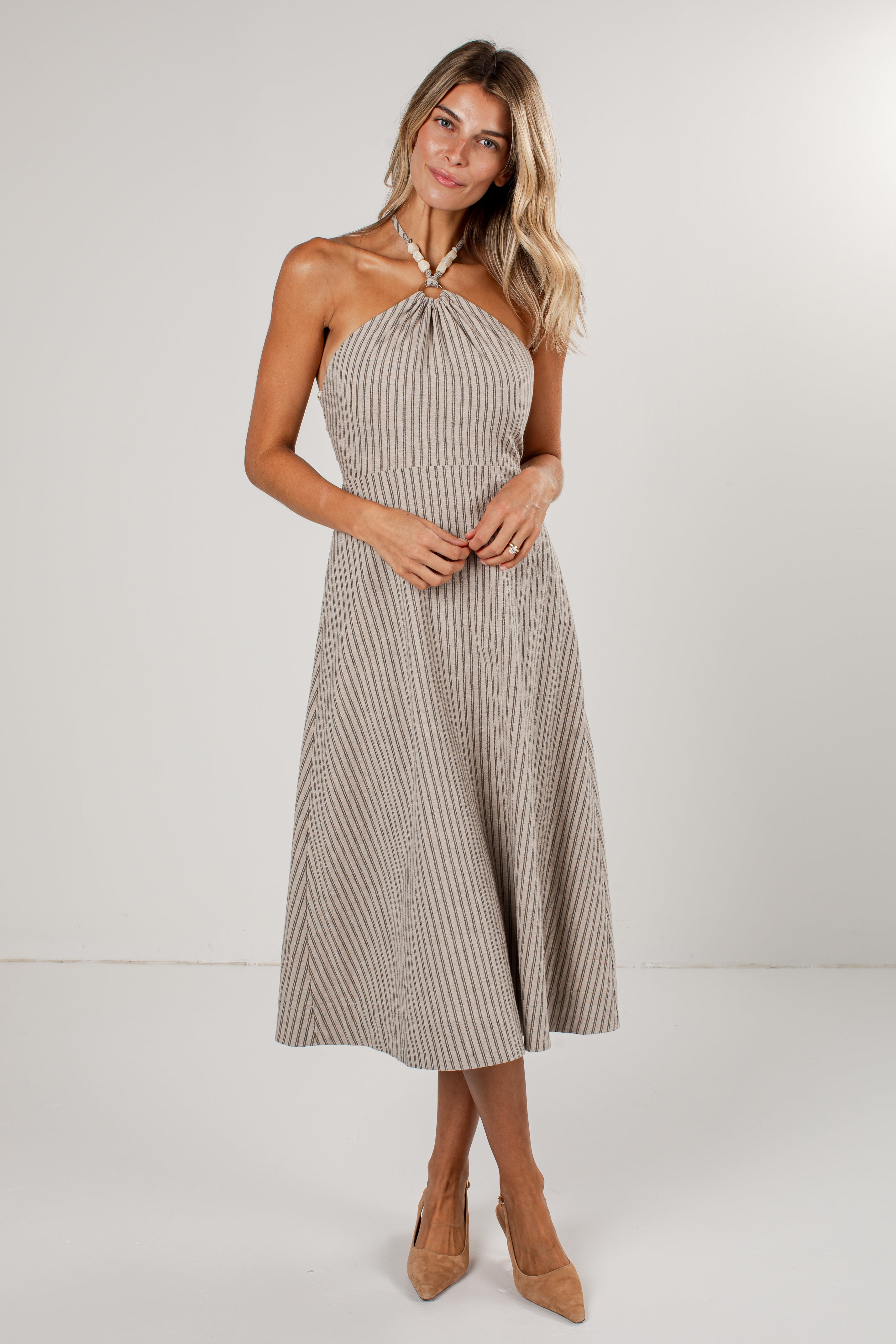 A woman modeling a halter-neck midi dress with vertical pinstripes in beige and white, paired with tan pointed-toe heels against a plain light gray studio background.