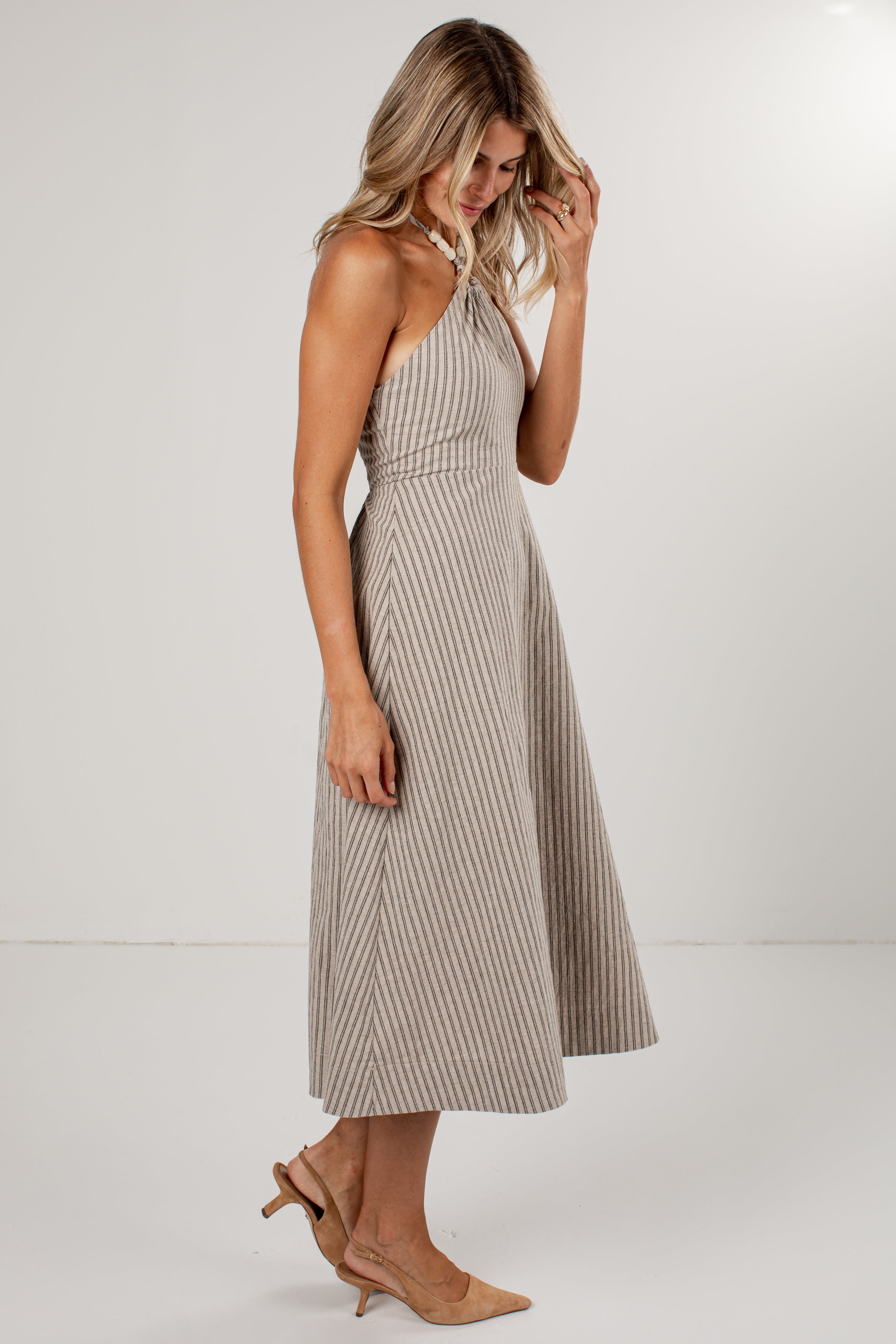 A woman modeling a sleeveless halter midi dress with a beige and gray vertical stripe pattern, paired with beige slingback heels, set against a plain light gray studio background.
