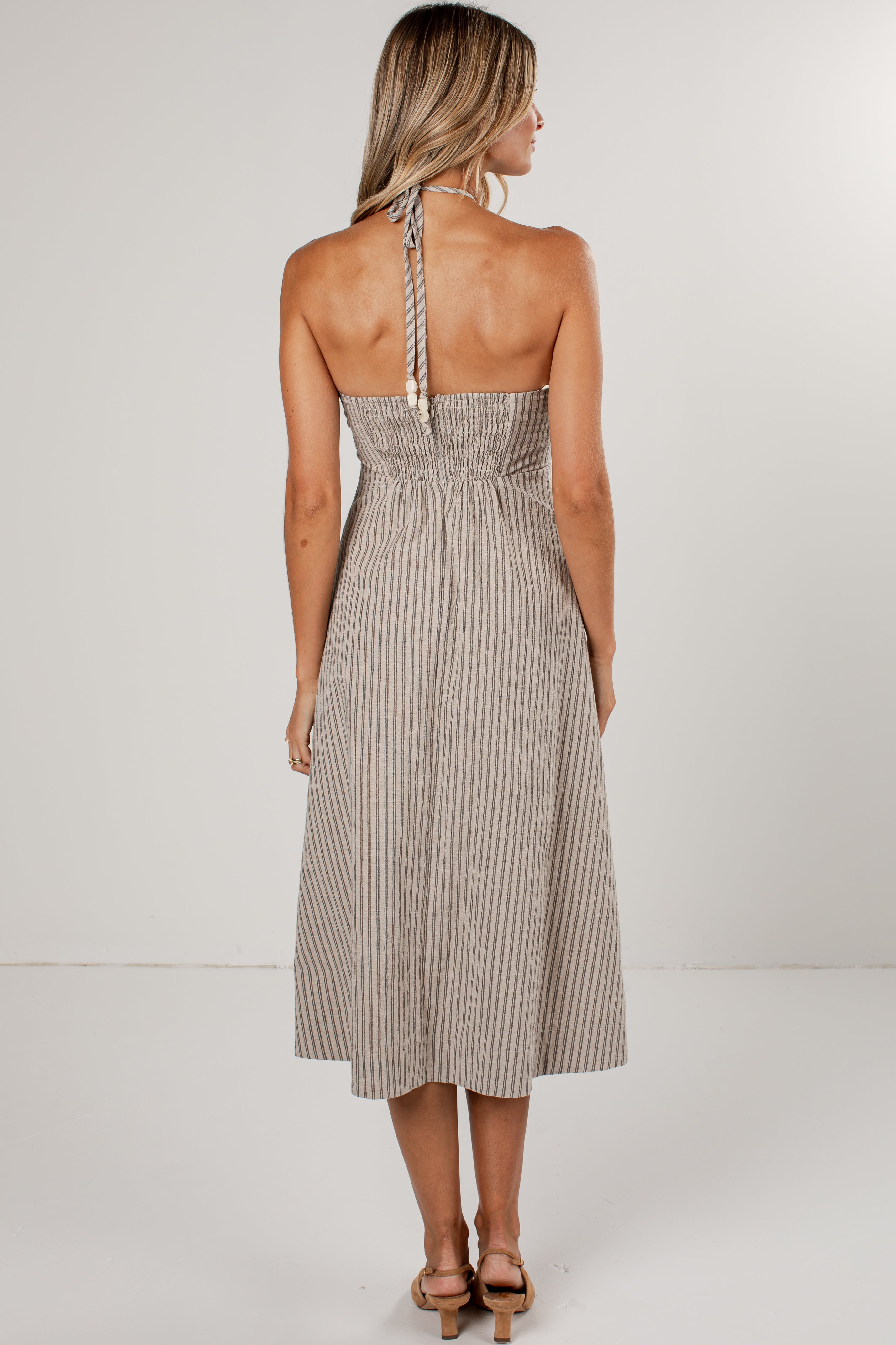 A woman viewed from the back wearing a sleeveless halter midi dress with thin vertical pinstripes in beige and white, paired with tan heeled sandals against a plain light gray studio background.
