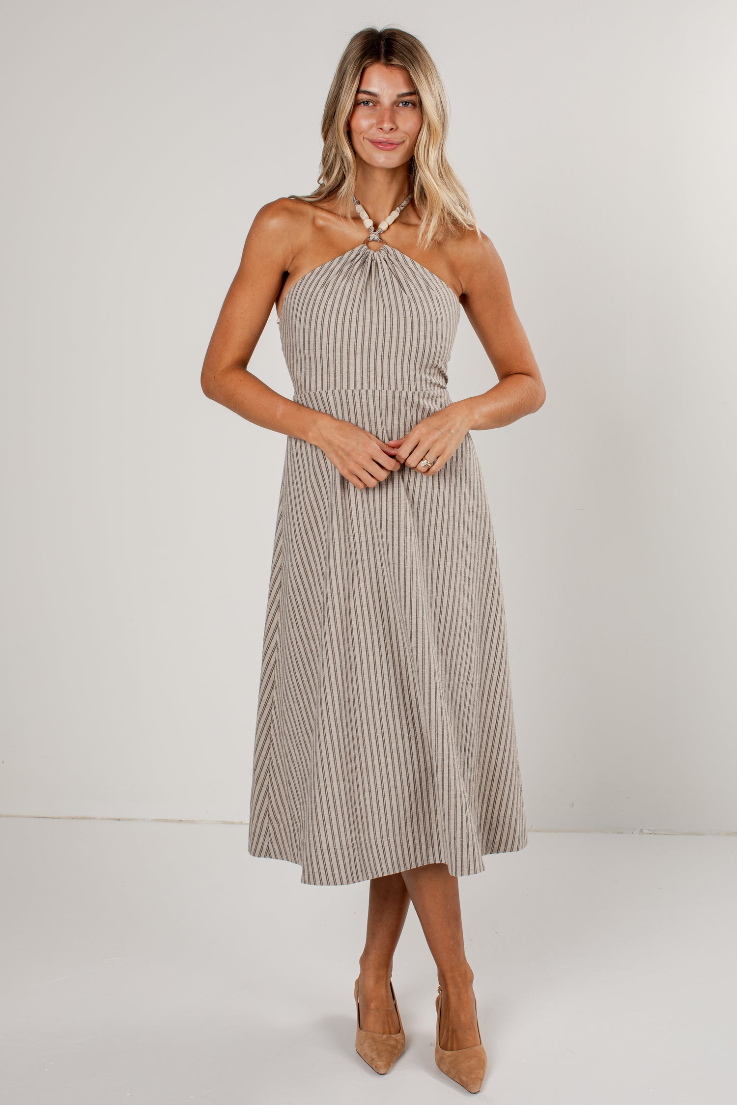 A woman modeling a sleeveless halter midi dress with vertical pinstripes in beige and white, paired with tan high heels, against a plain light gray studio background.