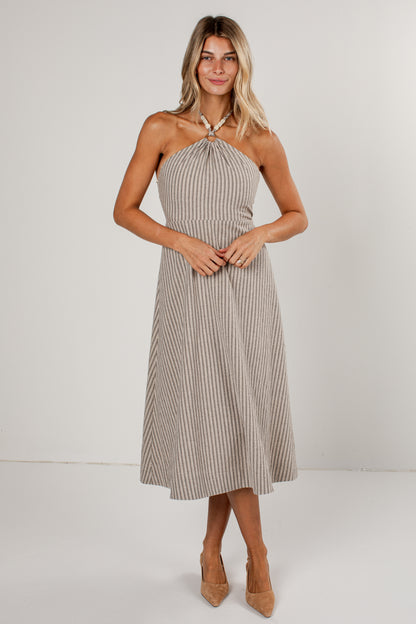 A woman modeling a sleeveless halter midi dress with vertical pinstripes in beige and white, paired with tan high heels, against a plain light gray studio background.