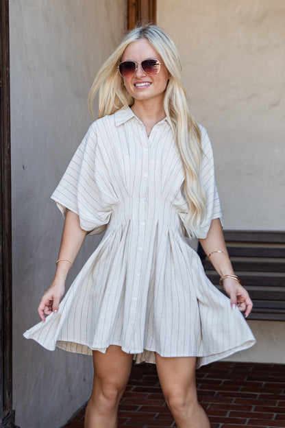A woman with long blonde hair wearing a cream and white vertically striped short-sleeve button-up dress, sunglasses, and bracelets, standing in front of a stucco wall.