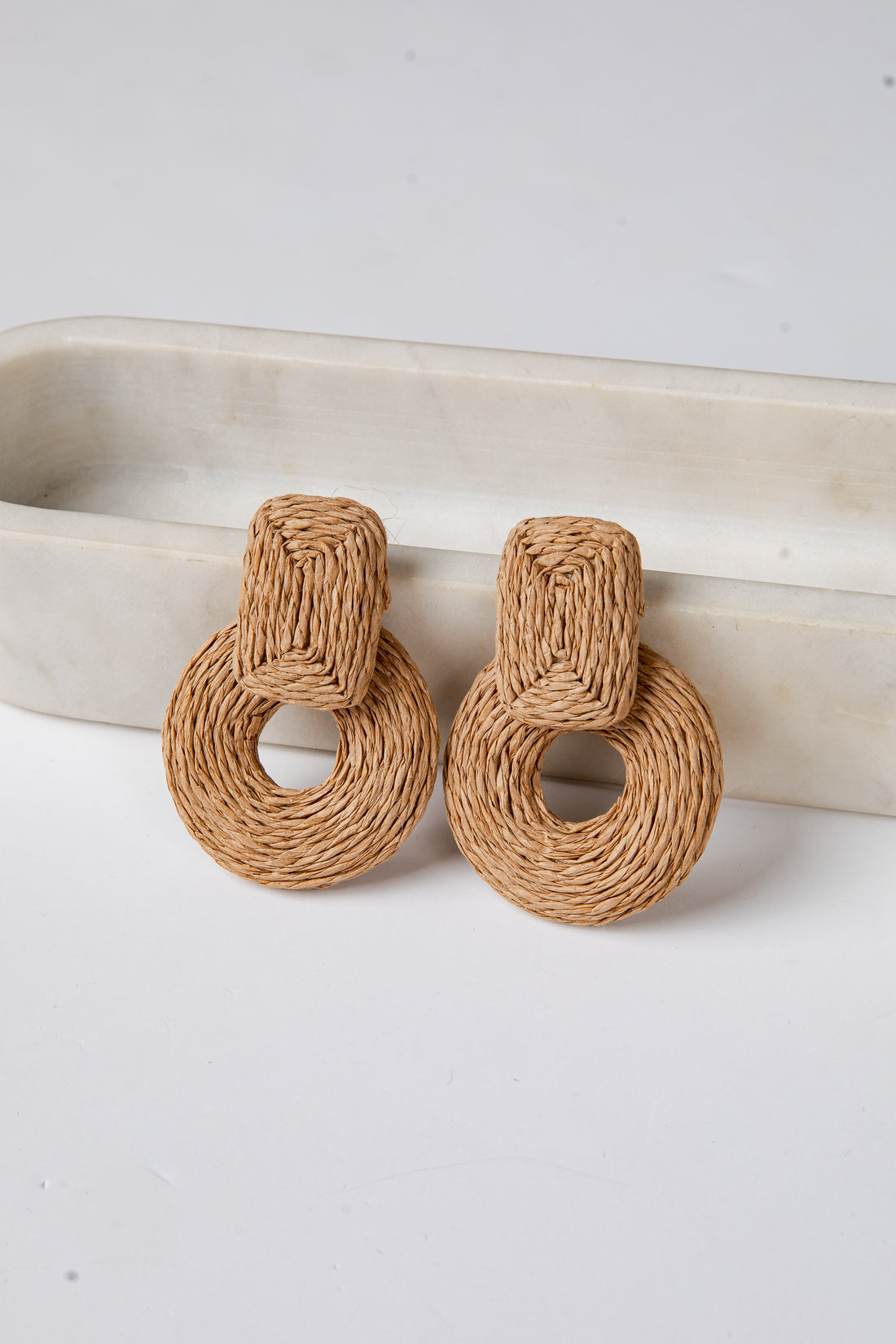 Pair of brown woven earrings on a white surface with a beige dish in the background.