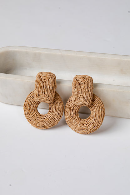 Pair of brown woven earrings on a white surface with a beige dish in the background.