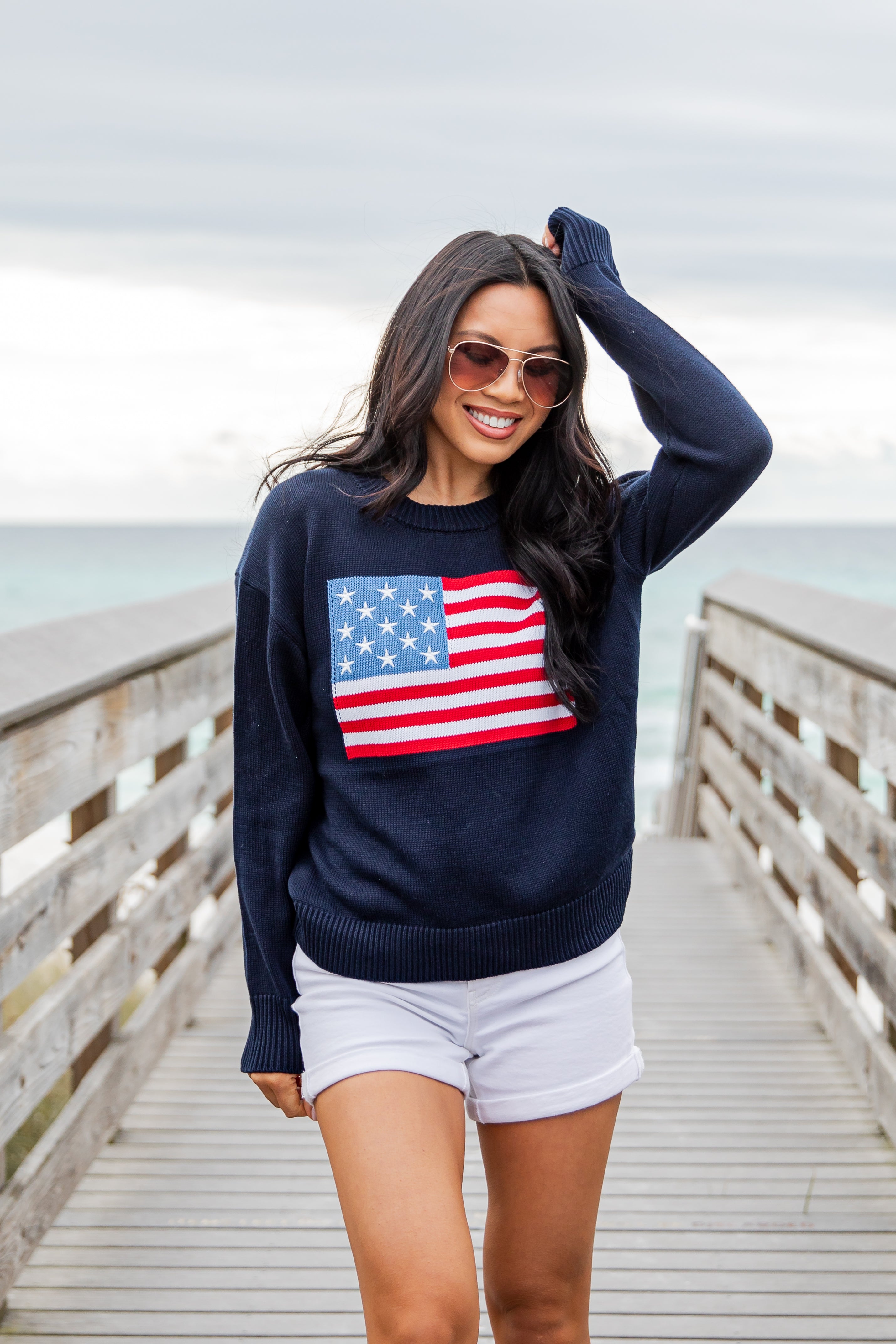 Woman wearing a navy sweater with an American flag design on a wooden pier by the ocean.