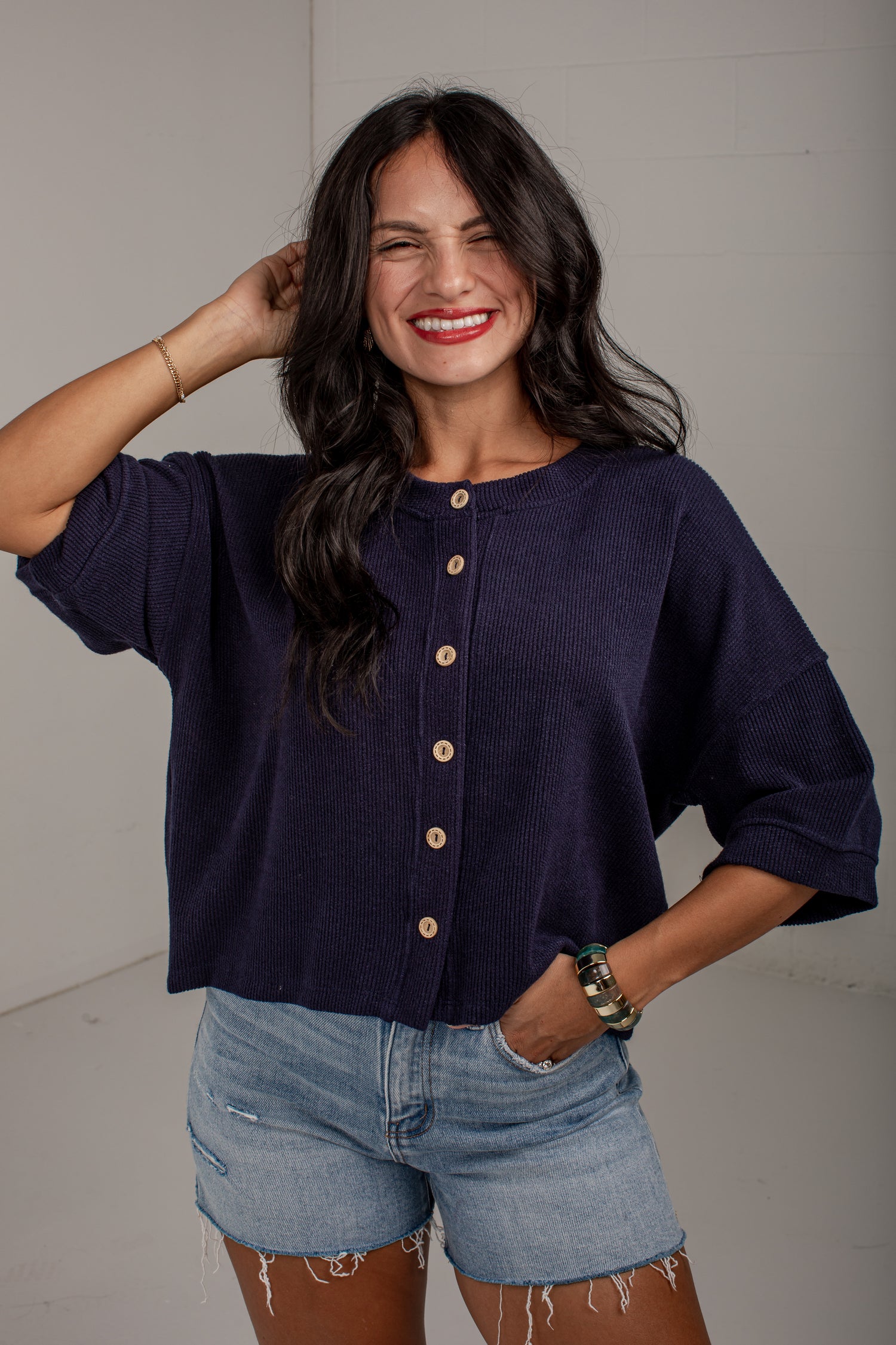 A woman wearing a dark navy blue short-sleeve button-up top with light wood buttons and light-wash denim shorts, posing against a light gray wall.