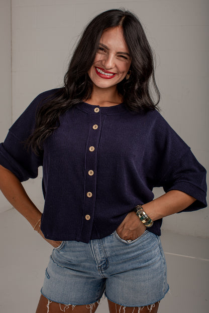 A woman wearing a dark navy blue ribbed knit cardigan with gold buttons and light-wash denim shorts, posing against a neutral light gray wall.