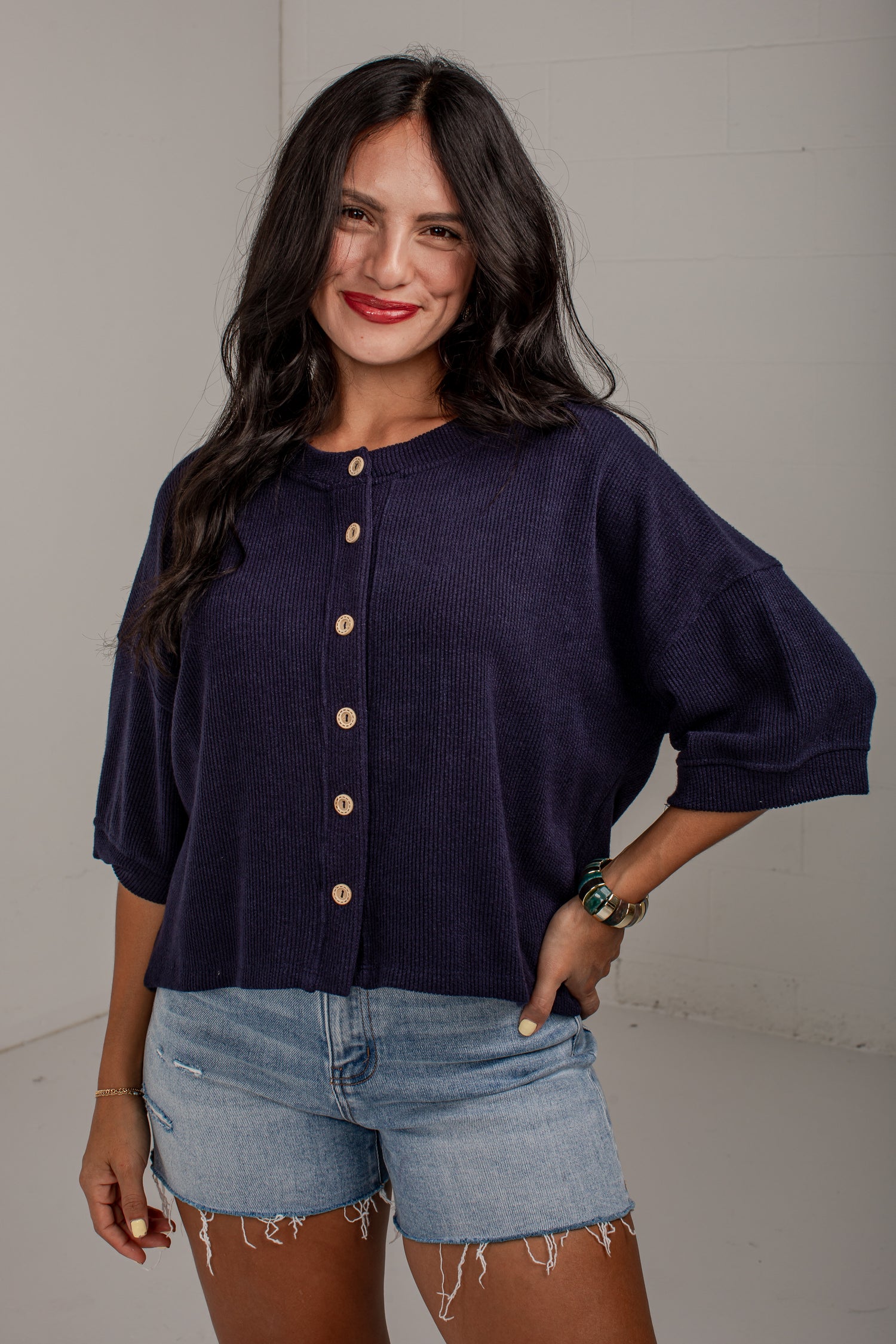 A woman modeling a dark navy blue short-sleeve button-up top with light brown buttons, paired with light-wash denim shorts, against a neutral light gray background.