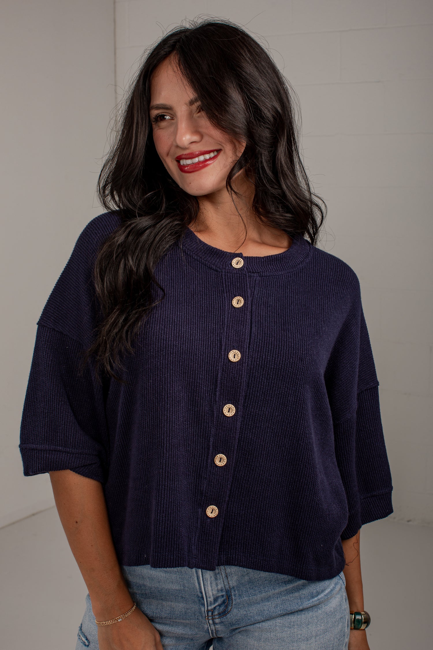 A woman wearing a dark navy ribbed knit cardigan with a button-down front and light-colored buttons, paired with light blue jeans, standing against a light gray wall.