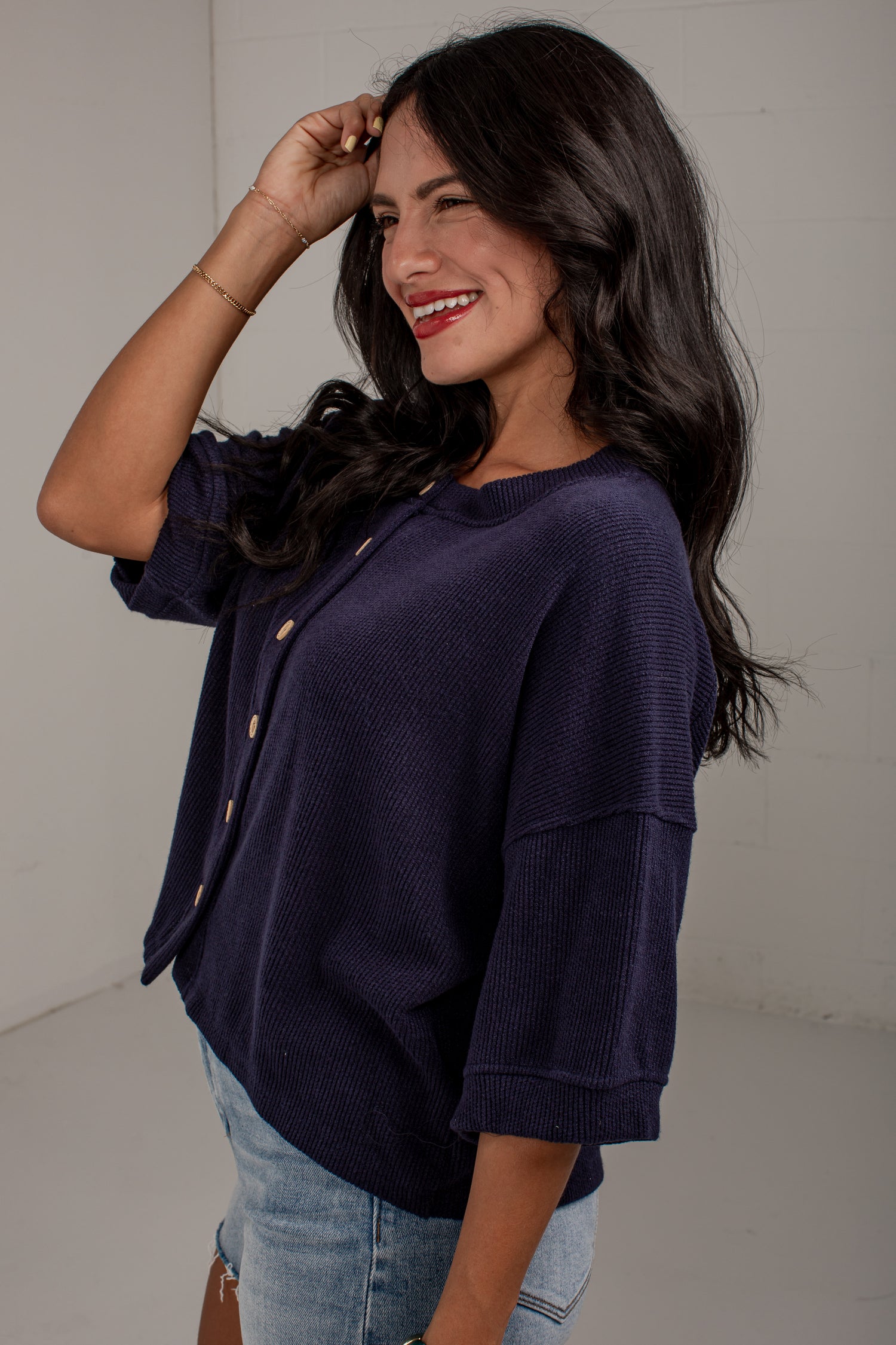 A woman wearing a dark purple ribbed knit button-up top with rolled three-quarter sleeves and light blue jeans, posing in a studio setting.