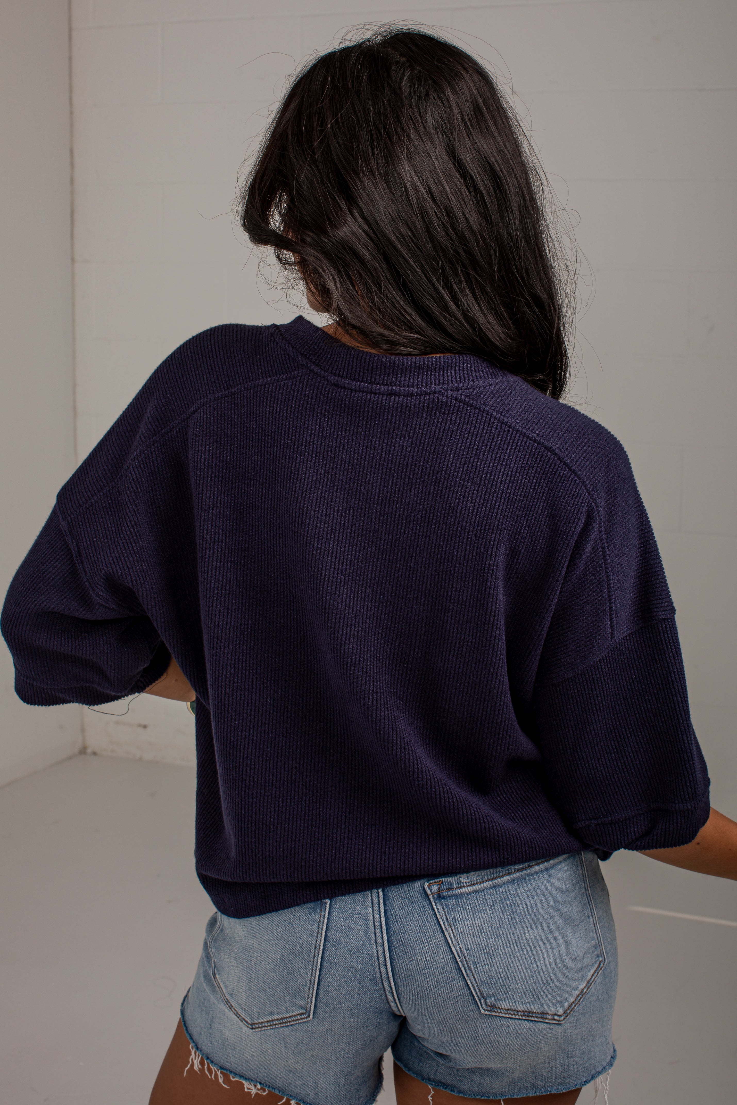 A person shown from behind wearing a dark navy blue cropped ribbed sweatshirt and light blue denim shorts in a plain indoor setting.