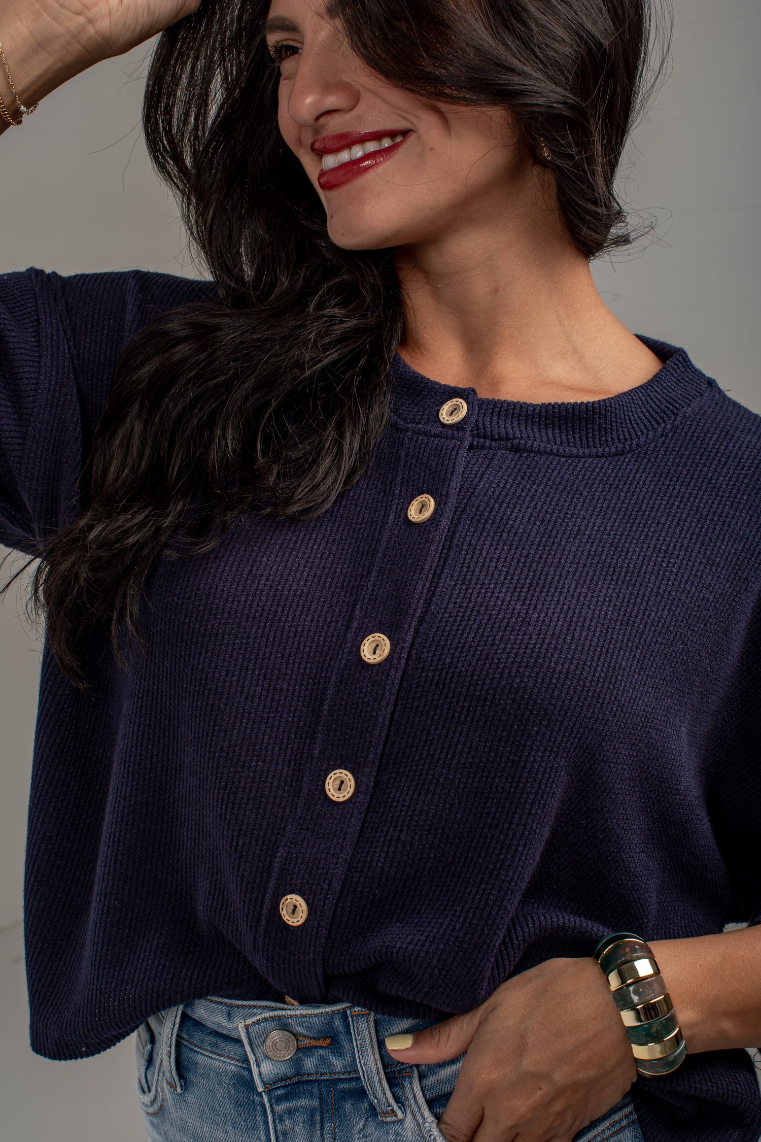 A woman wearing a navy blue ribbed knit top with a buttoned front, paired with blue jeans, shown in a close-up shot against a neutral gray background.