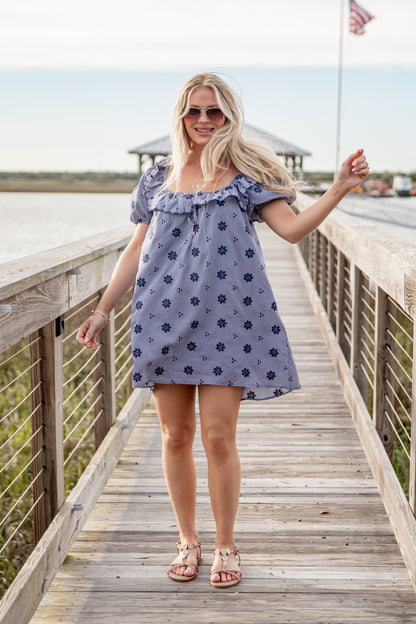 Woman in a blue floral dress standing on a wooden boardwalk by a waterfront.