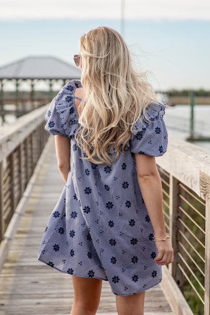 Woman in a blue floral dress standing on a wooden dock by a body of water.