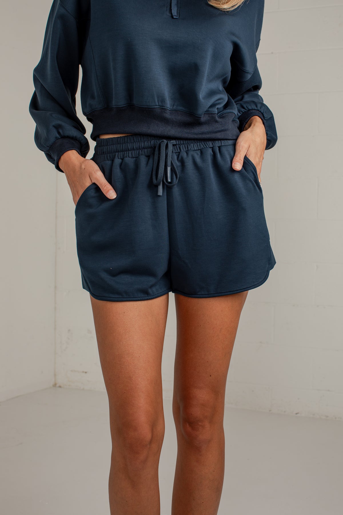 A person wearing a matching navy blue lounge set, featuring a cropped long-sleeve top and coordinating shorts with a drawstring waist and side pockets, standing against a plain off-white wall.