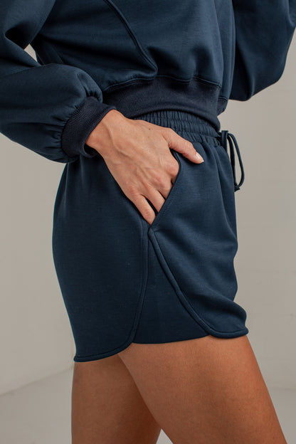Close-up of a person wearing dark navy sweat shorts with a side pocket and drawstring waist, paired with a matching navy sweatshirt.