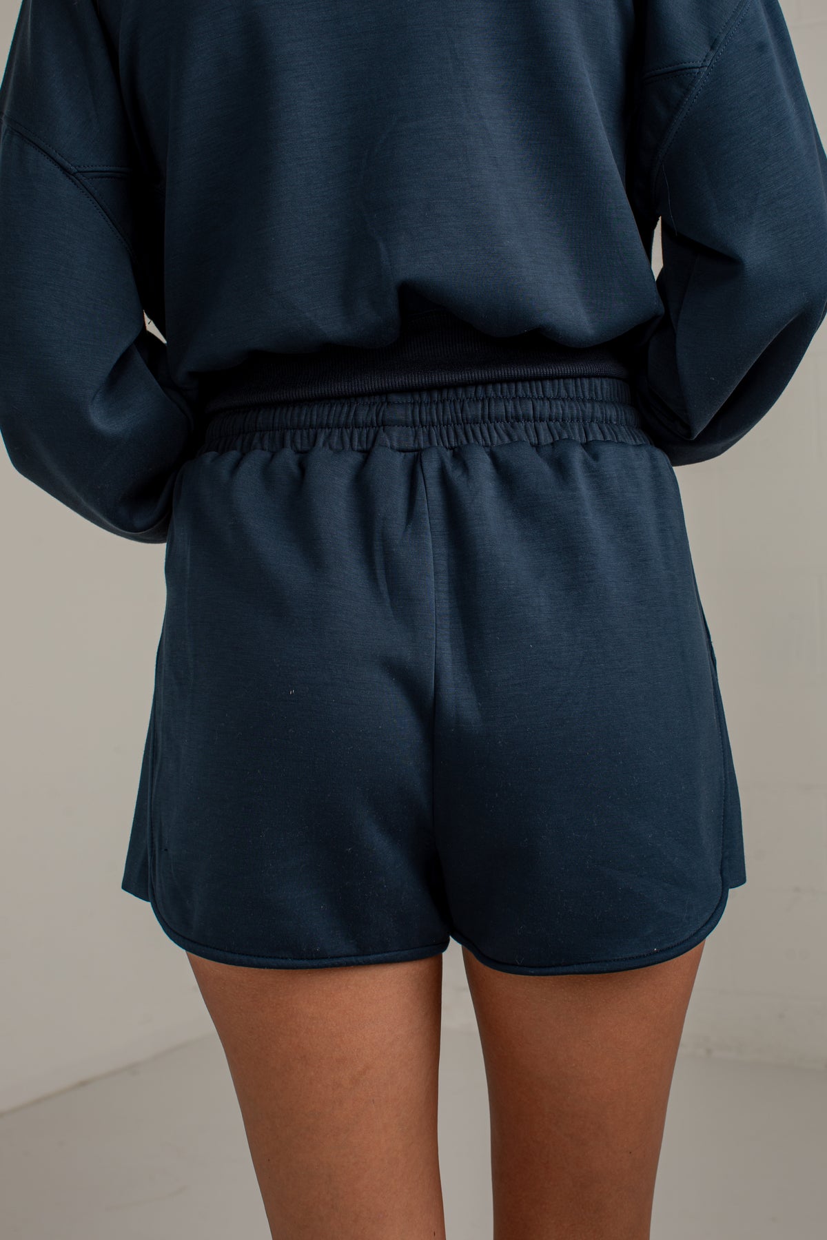 A person wearing dark navy blue sweat shorts and a matching sweatshirt, shown from the back against a plain light background.
