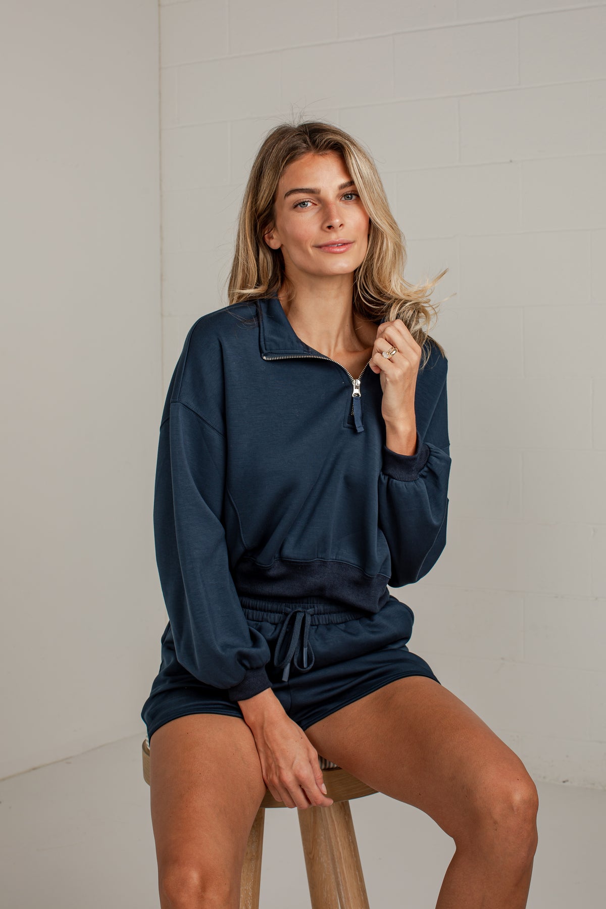 A woman sits on a wooden stool wearing a matching navy blue lounge set consisting of a quarter-zip sweatshirt and shorts, set against a white brick wall.