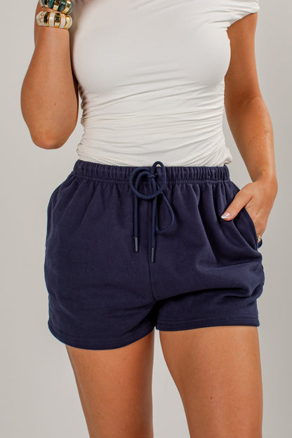 A person wearing navy blue drawstring shorts and a white top, shown from the chest to the upper thighs against a light gray studio background.
