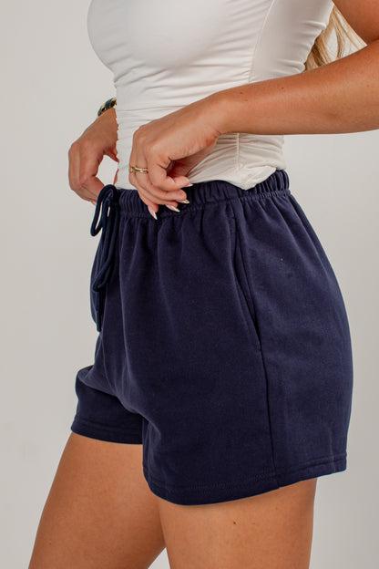 A close-up of a person wearing navy blue drawstring shorts and a white ribbed top, standing against a light gray studio background.