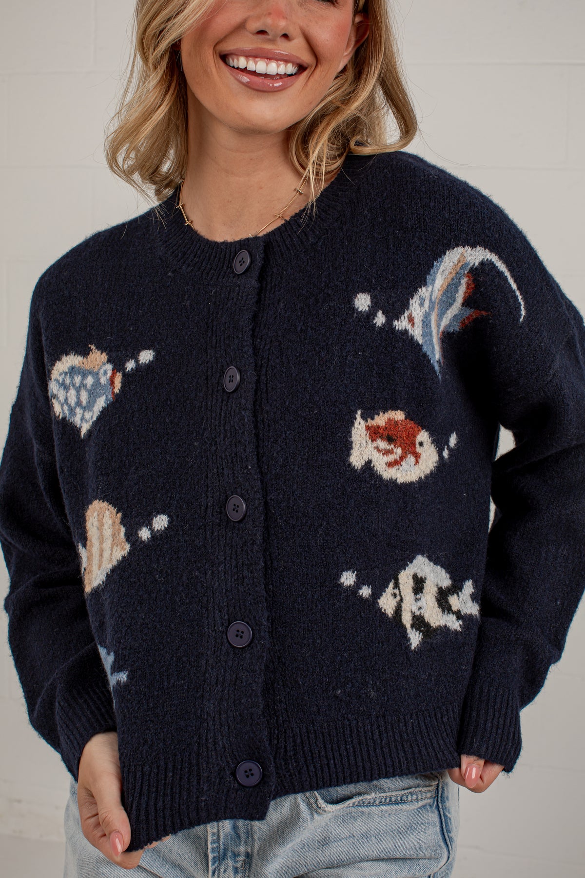 A woman wearing a dark navy blue knit cardigan with knitted seashell and shell-like motifs in cream, light blue, and rust red, paired with light-wash jeans against a white brick wall.