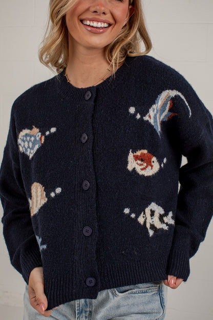 A woman wearing a dark navy blue knit cardigan with knitted seashell and shell-like motifs in cream, light blue, and rust red, paired with light-wash jeans against a white brick wall.