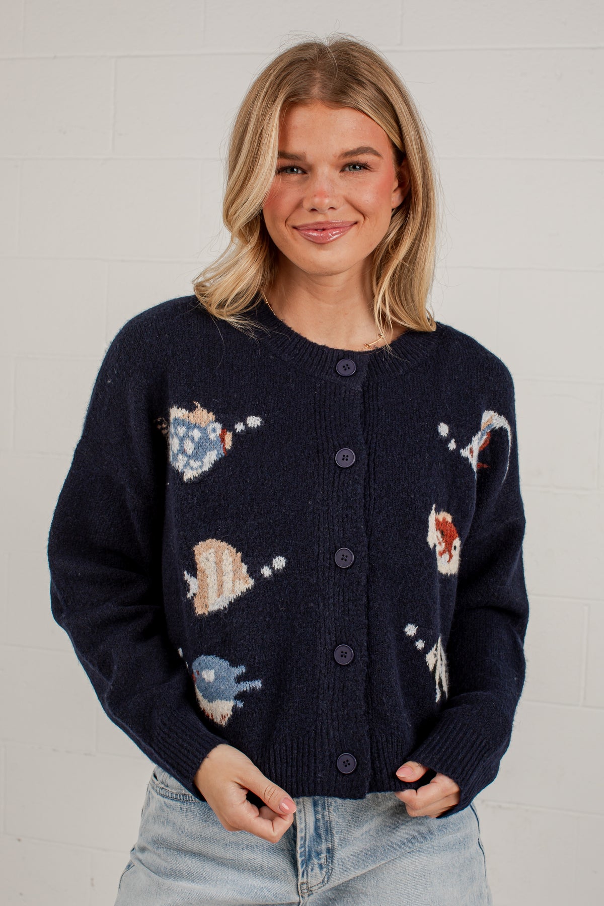 A woman wearing a navy blue knit cardigan with colorful fish and sea-creature motifs, paired with light-wash jeans, standing against a white brick wall.