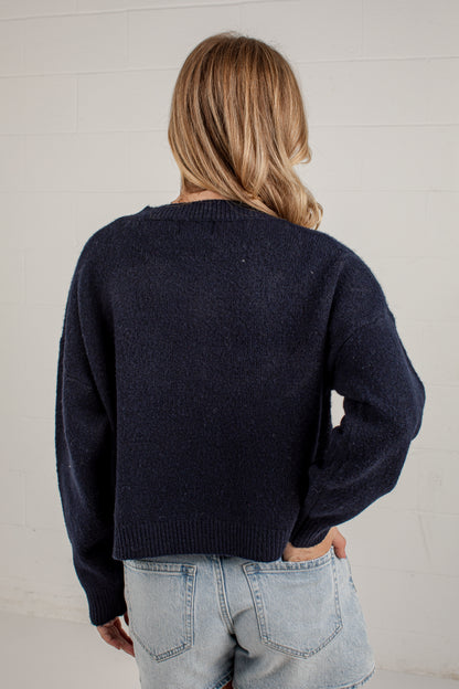 A woman viewed from behind wearing a dark navy blue knit sweater and light-wash denim shorts against a white brick wall.