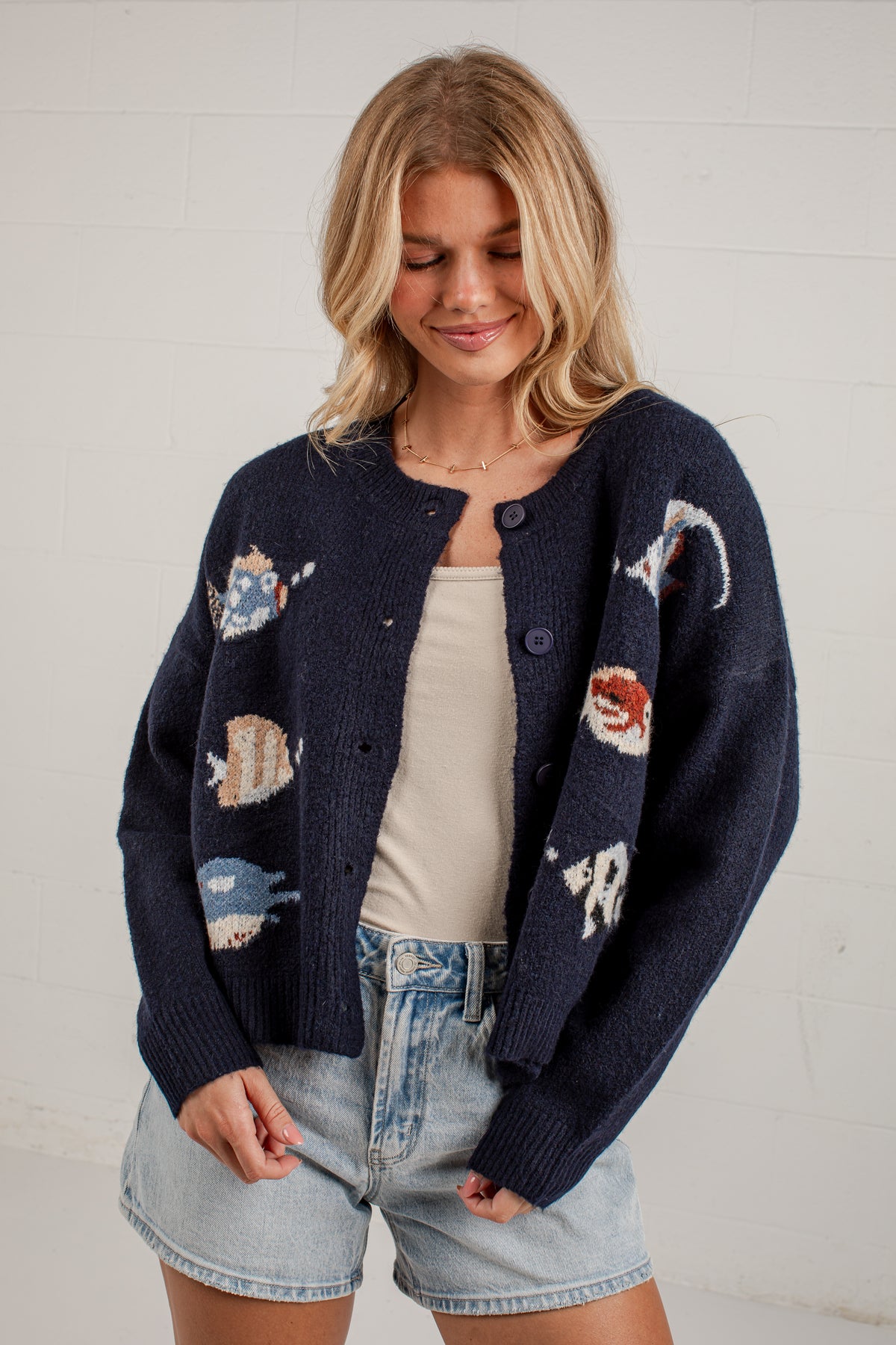 A woman wearing a navy blue knit cardigan with colorful fish motifs, styled over a cream tank top and light-wash denim shorts against a white brick wall.