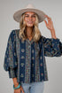 A woman wearing a navy blue bohemian-style blouse with vertical floral patterns, paired with a wide-brimmed beige hat and light blue jeans, photographed against a solid gray studio background.