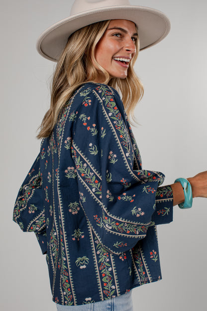 A woman wearing a navy blue bohemian-style embroidered blouse with floral patterns, a wide-brimmed beige hat, and light blue jeans, photographed against a neutral gray studio background.