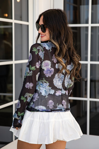 Woman wearing a floral blouse and white skirt in front of a window.