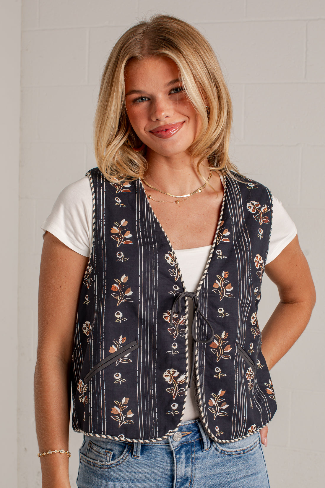 A woman wearing a navy blue vest with a floral and striped pattern over a white t-shirt and blue jeans, standing against a light gray brick wall.