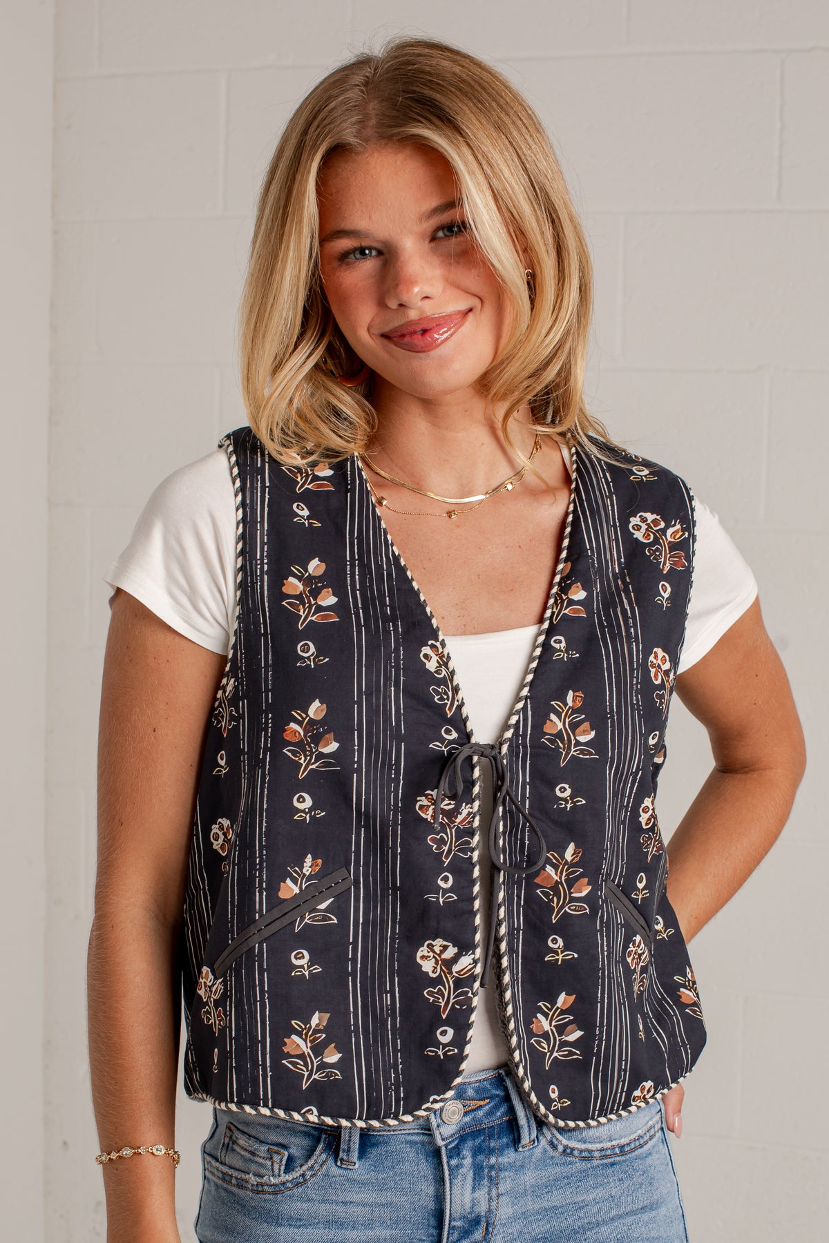 A woman wearing a navy blue vest with a floral and striped pattern over a white t-shirt and blue jeans, standing against a light gray brick wall.