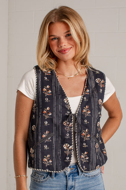 A woman wearing a navy blue vest with a floral and striped pattern over a white t-shirt and blue jeans, standing against a light gray brick wall.