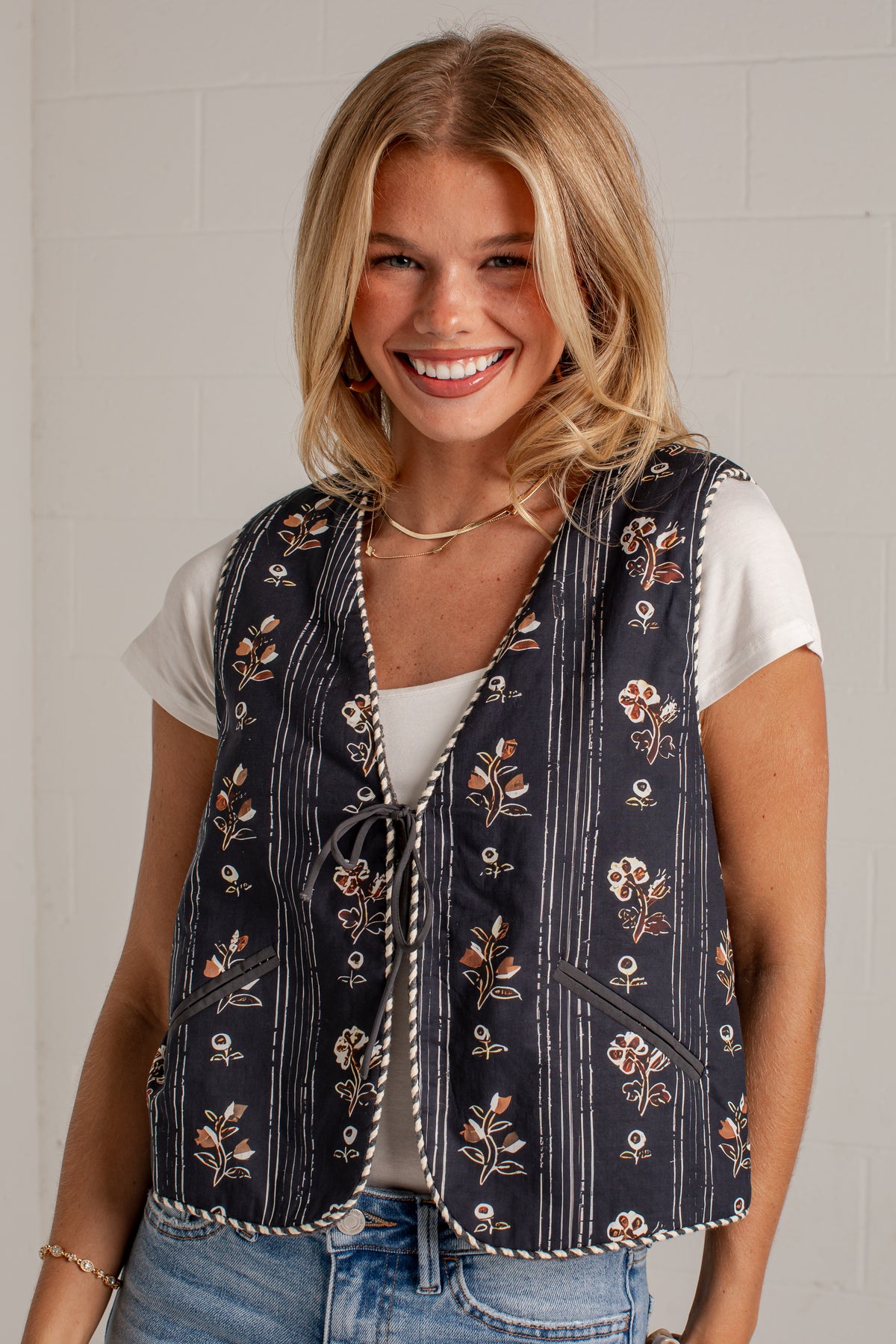 A smiling blonde woman wearing a navy blue floral vest over a white t-shirt and blue jeans, standing against a white brick wall.