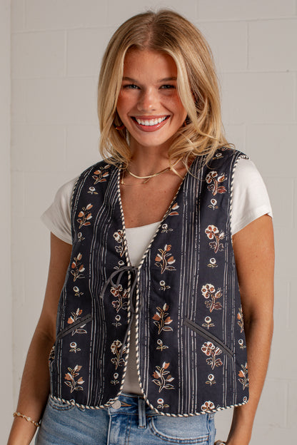 A smiling blonde woman wearing a navy blue floral vest over a white t-shirt and blue jeans, standing against a white brick wall.