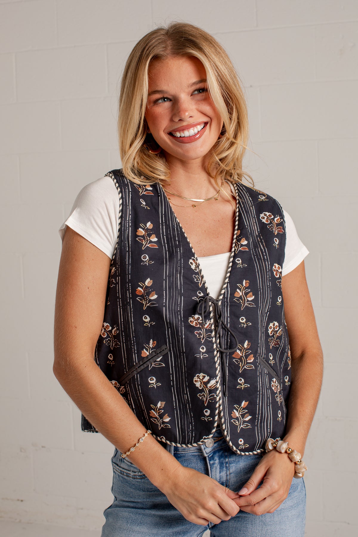 A smiling blonde woman wearing a sleeveless navy vest with a white floral print, a white t-shirt, and blue jeans, standing against a light gray brick wall.