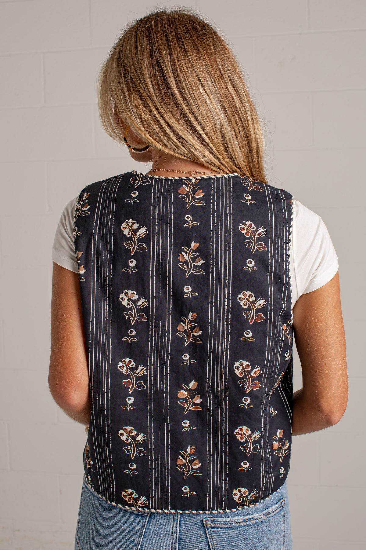 A woman viewed from behind wearing a sleeveless navy vest with vertical white stripes and a floral pattern, layered over a white t-shirt and blue jeans.