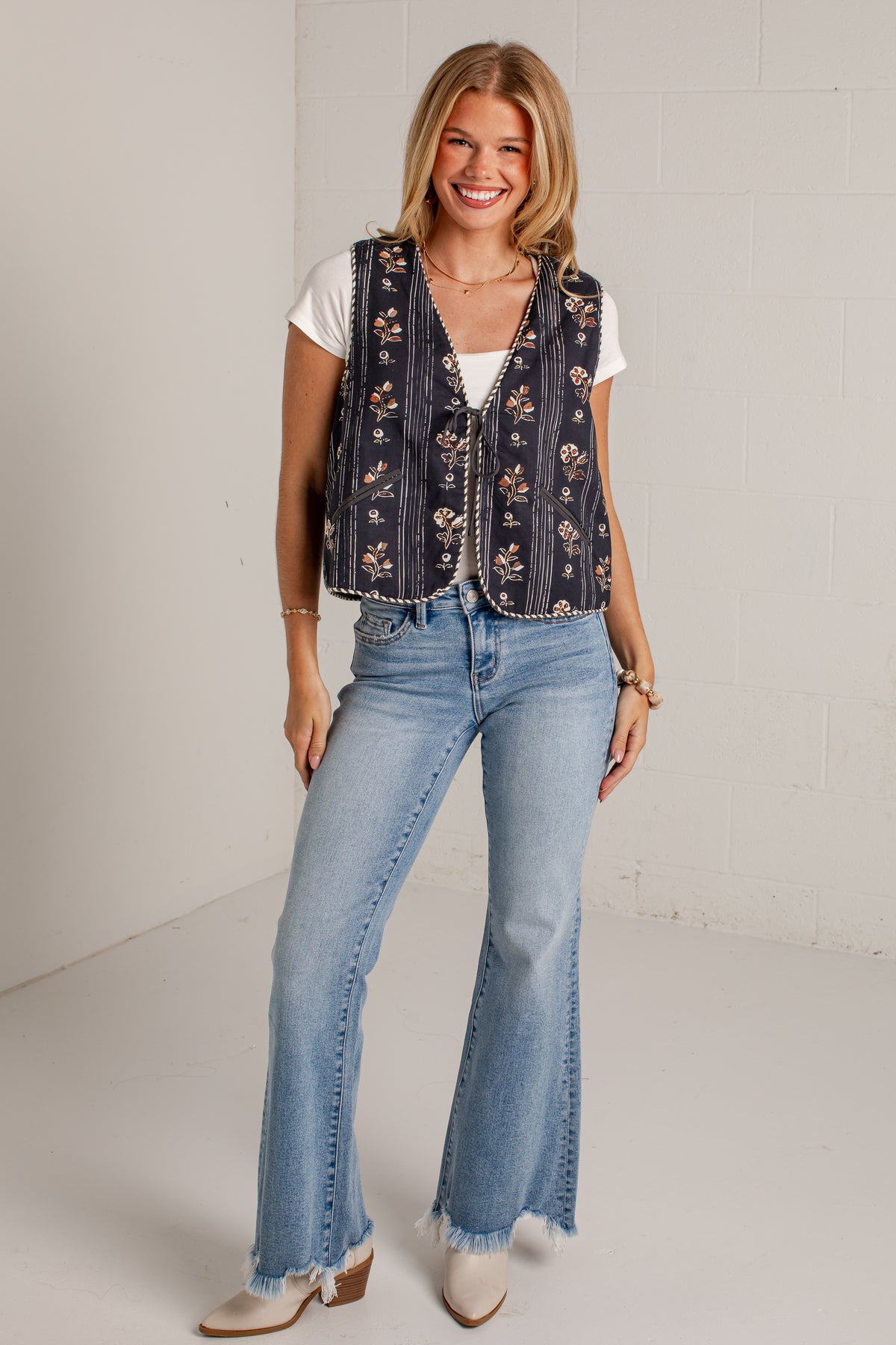 A woman modeling a sleeveless navy blue embroidered vest with floral patterns over a white t-shirt and light-wash flare jeans, standing against a white brick wall.