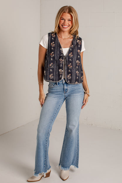 A woman modeling a sleeveless navy blue embroidered vest with floral patterns over a white t-shirt and light-wash flare jeans, standing against a white brick wall.