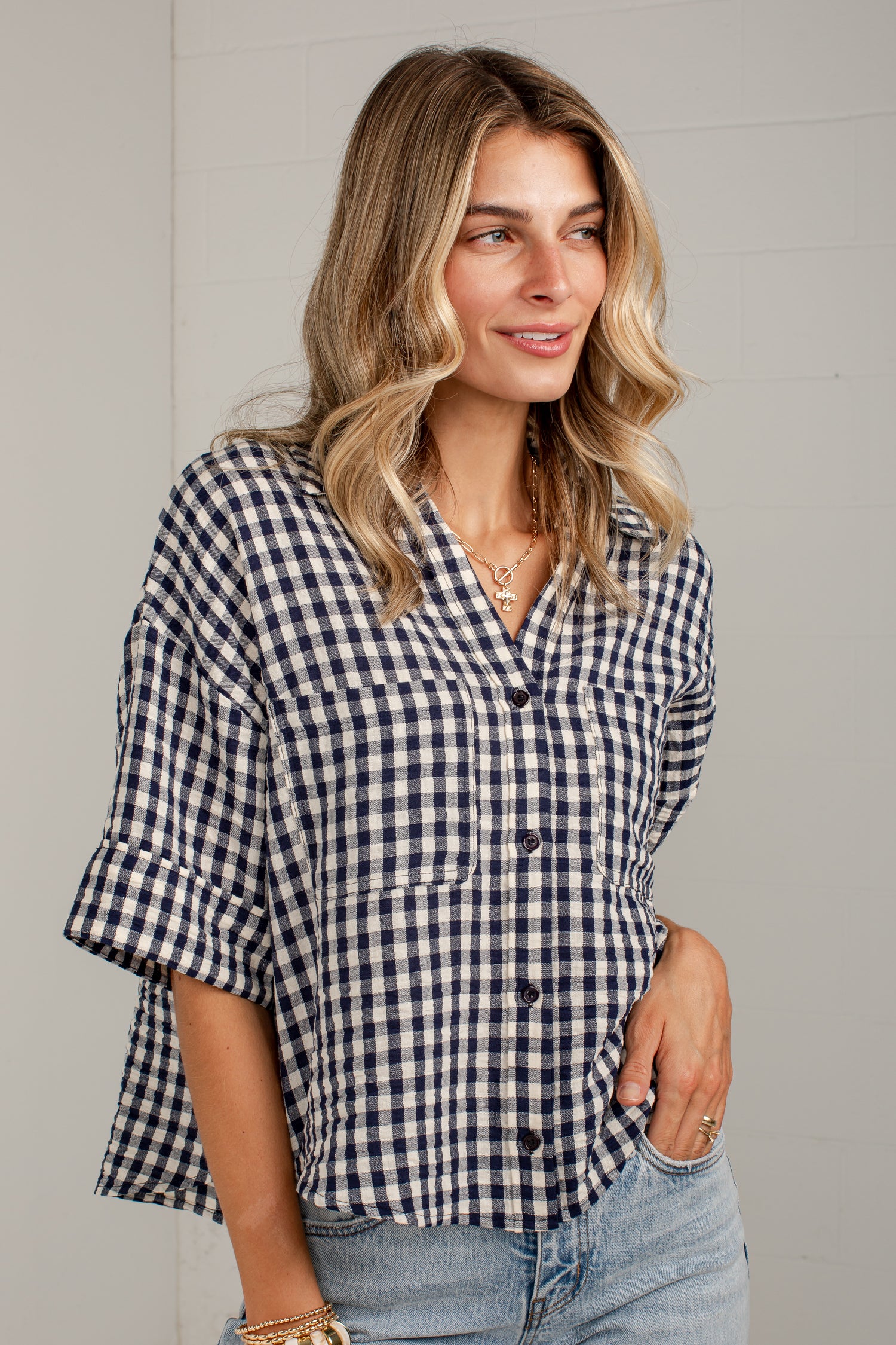 A woman wearing a navy and white gingham button-down shirt with rolled sleeves and light-wash jeans, standing against a light gray paneled wall.