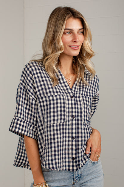 A woman wearing a navy and white gingham button-down shirt with rolled sleeves and light-wash jeans, standing against a light gray paneled wall.