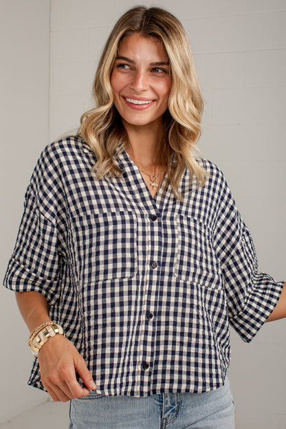 A woman wearing a navy and white gingham button-up shirt with three-quarter sleeves, paired with light blue jeans, standing against a light gray paneled wall.